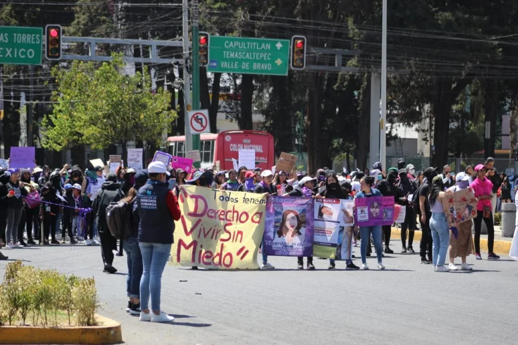 “Manifestación en una calle urbana con un grupo numeroso de personas sosteniendo pancartas y carteles; al frente destaca una lona con el mensaje ‘Todos tenemos derecho a vivir sin miedo’, mientras semáforos y señalamientos viales aparecen en el fondo.”