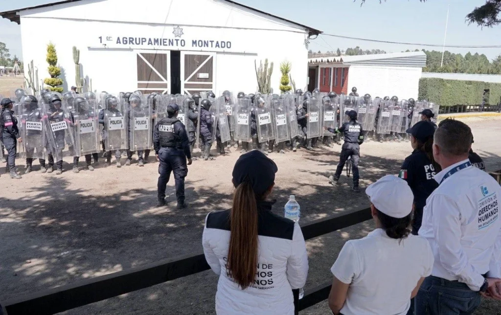 “Formación de elementos de policía estatal con escudos transparentes alineados frente a un edificio rotulado ‘1° Agrupamiento Montado’; en primer plano, varias personas observan la práctica o entrenamiento desde fuera del área delimitada.”
