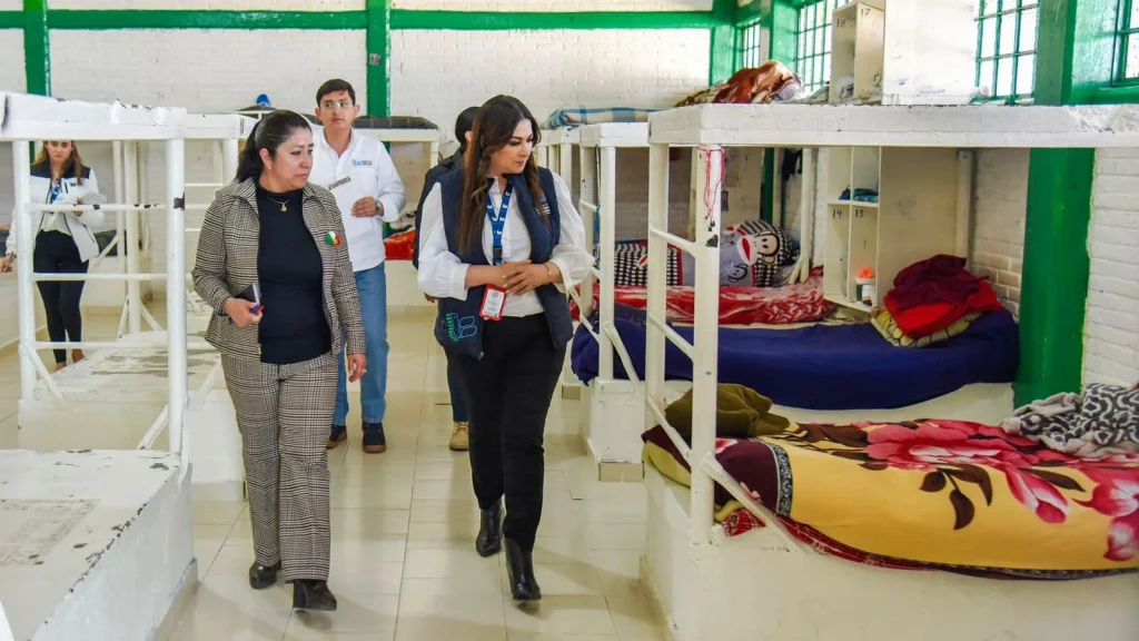 “Área interior con literas metálicas dispuestas en filas, donde varias personas recorren el espacio realizando una inspección; se observan camas con cobijas y pertenencias en un entorno con paredes blancas y estructuras pintadas en verde.”