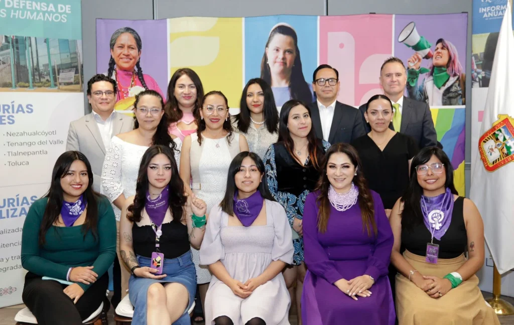 “Grupo numeroso de personas posando para una fotografía oficial en un espacio interior, con varias filas de pie y sentadas; al fondo hay un panel colorido con retratos y un mensaje institucional relacionado con derechos humanos, además de una bandera a un costado.”