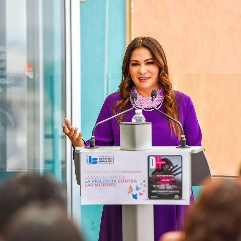 “Persona de pie detrás de un podio con dos micrófonos durante una presentación institucional; el podio muestra el logotipo de la Comisión de Derechos Humanos y el texto ‘La evolución de la violencia contra las mujeres’, en un espacio interior con paredes claras.”