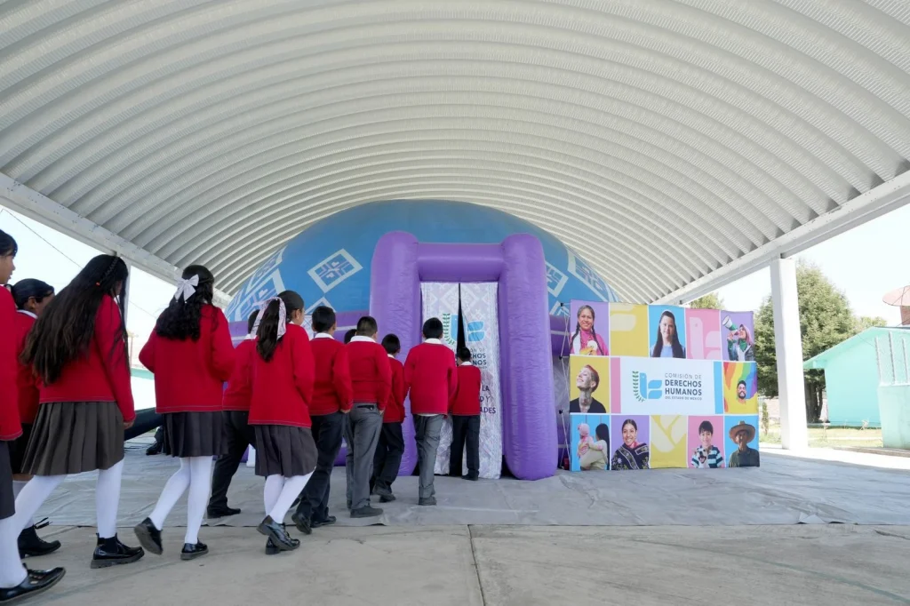 “Grupo de estudiantes formados en fila ingresando a una estructura inflable con forma de domo de color morado y azul, instalada bajo una techumbre; a un costado se observa un panel con el logotipo de la Comisión de Derechos Humanos y fotografías de diversas personas.”