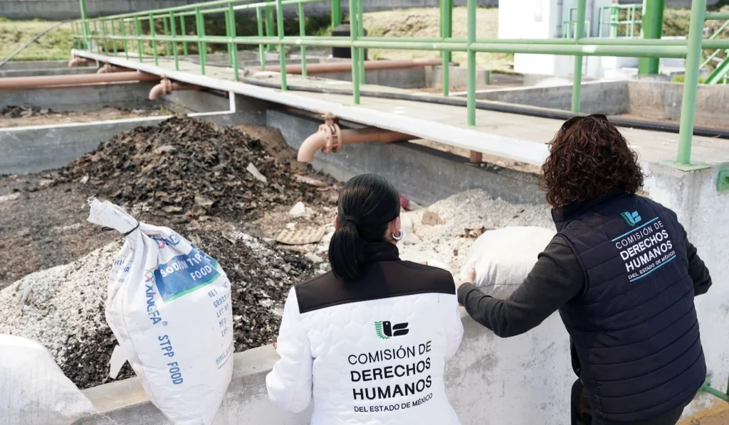 “Dos personas con chalecos de la Comisión de Derechos Humanos observan una zona industrial al aire libre con canales de concreto, tuberías y acumulaciones de residuos sólidos, como parte de una inspección técnica en instalaciones.”