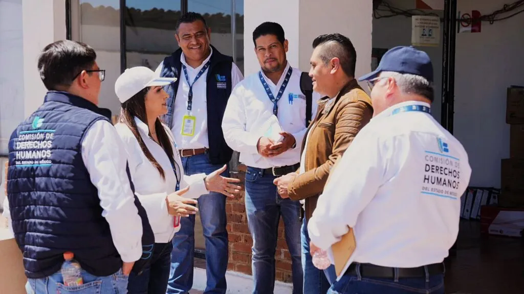 Grupo de seis personas reunidas en un espacio exterior frente a un edificio, conversando en círculo. Varias personas llevan chalecos y camisas con el logotipo de la Comisión de Derechos Humanos del Estado de México.