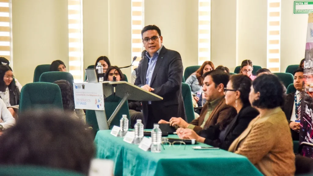 Persona de pie hablando desde un podio con micrófono en una sala de conferencias. Frente al podio hay una mesa con mantel verde donde varias personas están sentadas escuchando, con botellas de agua sobre la mesa