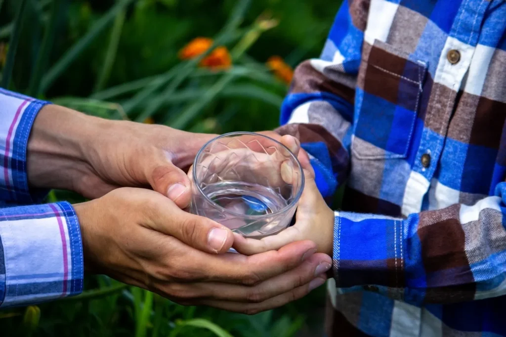 Primer plano de dos pares de manos sosteniendo juntas un vaso transparente con agua. Una persona entrega el vaso a otra más pequeña. Ambas personas llevan camisas de manga larga con estampado de cuadros en tonos azules y marrones. El fondo está desenfocado y muestra vegetación verde y flores naranjas, lo que sugiere un entorno exterior natural.