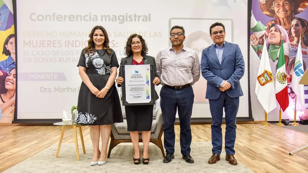 Cuatro personas de pie en un escenario durante un evento formal, posando frente a una pantalla de presentación que contiene el texto “Conferencia magistral”, “Derecho humano a la salud de las mujeres indígenas” y otras líneas parcialmente visibles.