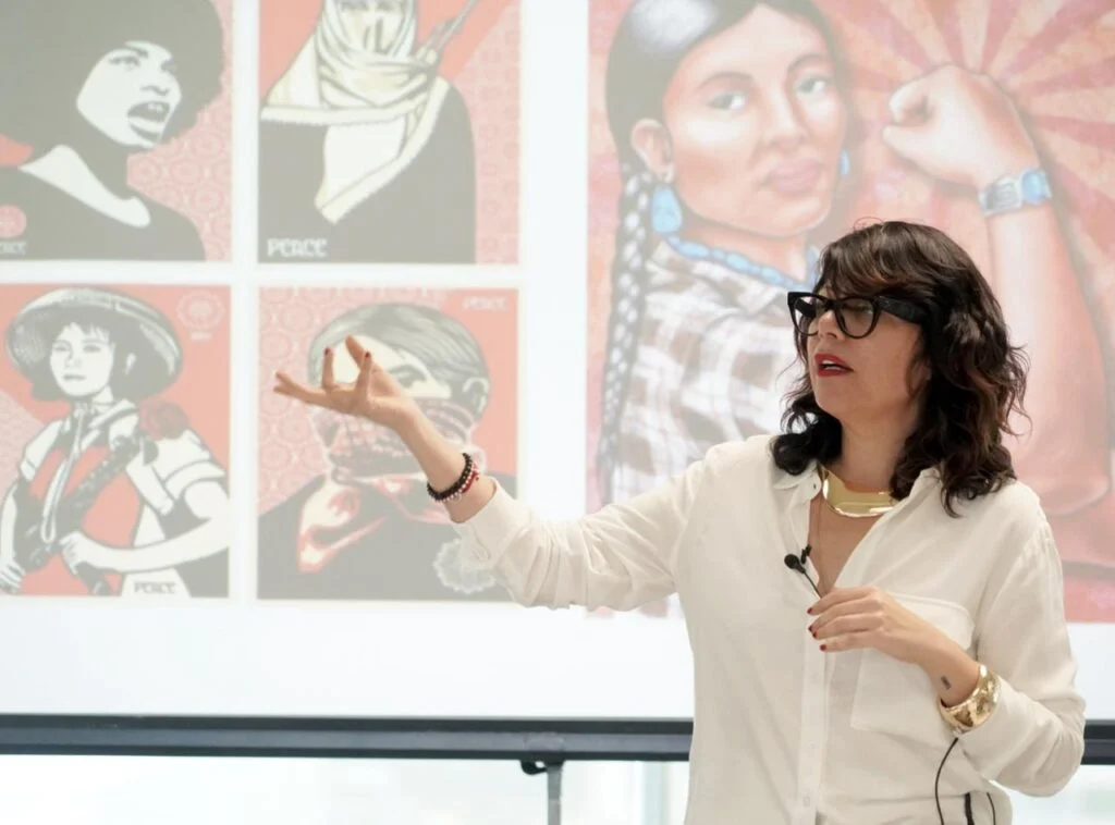 Persona de pie dando una presentación frente a una pantalla con ilustraciones gráficas de estilo cartel. La persona gesticula con una mano mientras expone, y viste ropa clara con accesorios visibles. La proyección incluye imágenes de figuras ilustradas en colores rojo y beige.