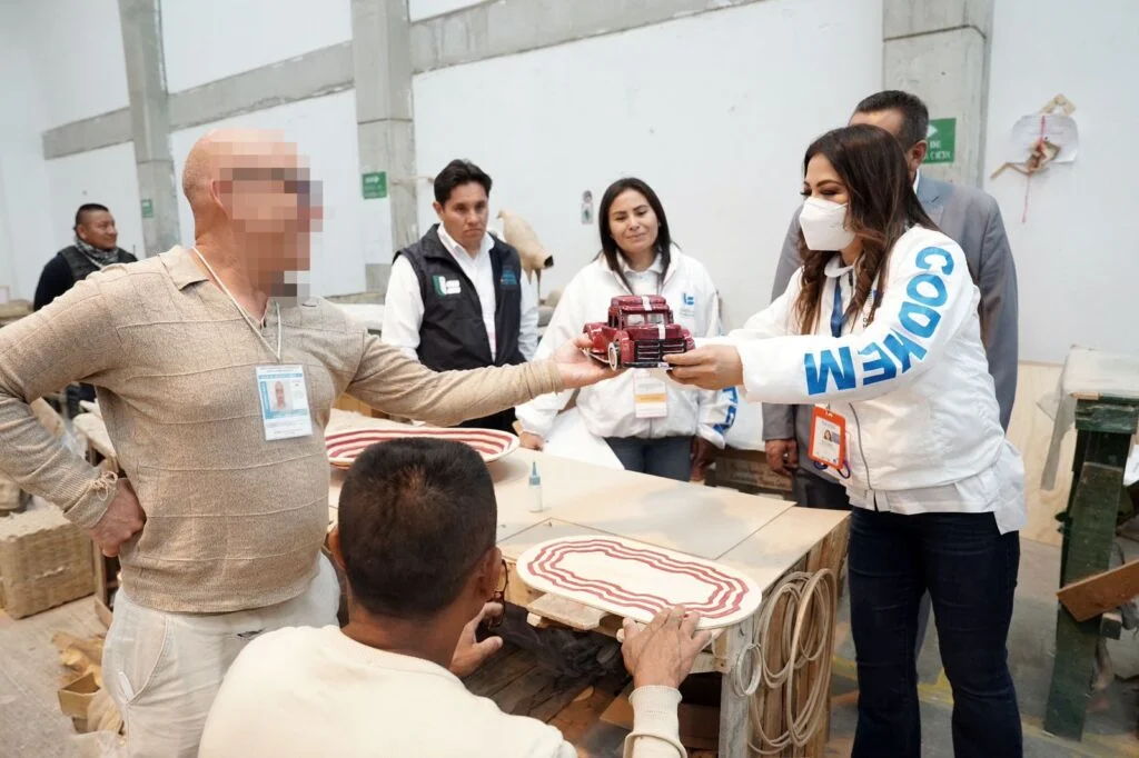 Varias personas dentro de un taller entregan una figura artesanal en forma de camión a otra persona de pie. La escena ocurre alrededor de una mesa de trabajo con piezas de madera y materiales en proceso. En el fondo se observan herramientas y más áreas de producción.
