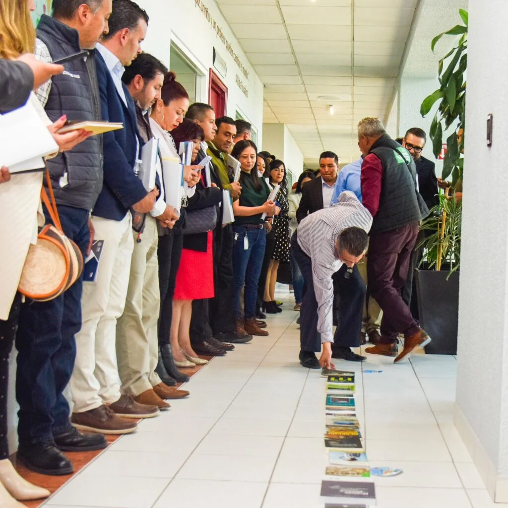 Grupo de personas formadas a ambos lados de un pasillo interior observa una dinámica en el suelo donde se colocan varios libros alineados. Una persona se inclina para acomodar un elemento mientras otras personas sostienen libretas y documentos. El espacio tiene piso de loseta clara, paredes blancas y plantas decorativas.