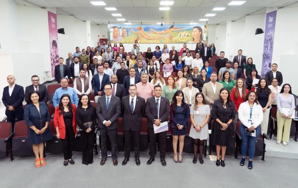 Fotografía grupal en un auditorio con decenas de personas organizadas en filas frente a asientos tipo teatro. Las personas posan de pie hacia la cámara, mientras al fondo se aprecia un mural colorido y banners institucionales en las paredes.