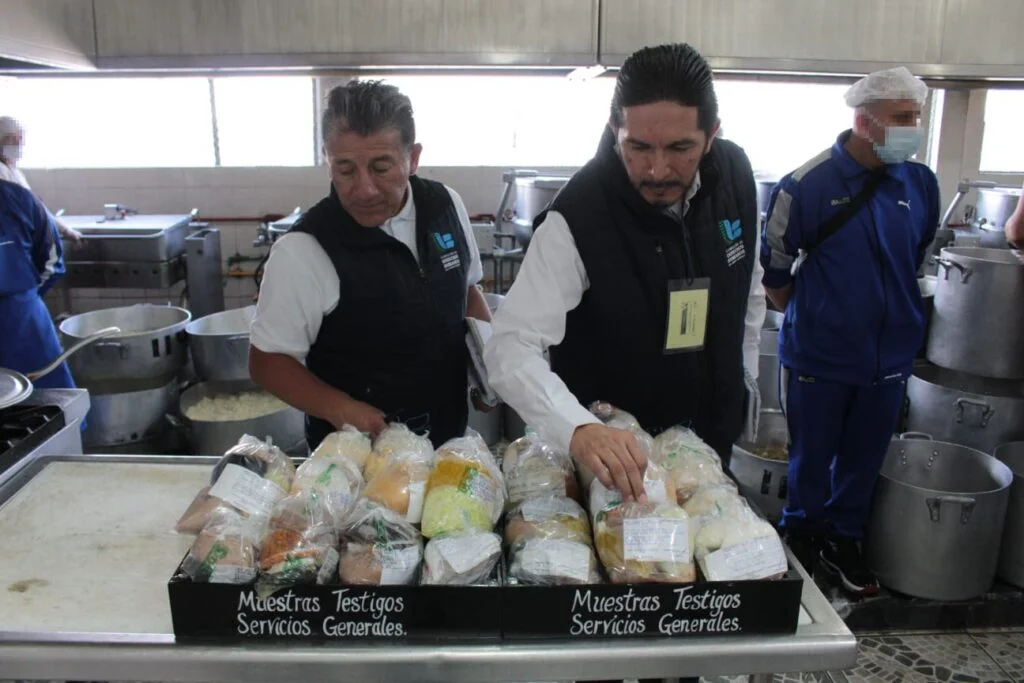 Cocina industrial donde varias personas revisan paquetes de alimentos colocados en bandejas metálicas. Los paquetes están etiquetados como “Muestras Testigos Servicios Generales”. Al fondo se observan ollas grandes, utensilios de cocina y personal con uniforme y equipo de protección.