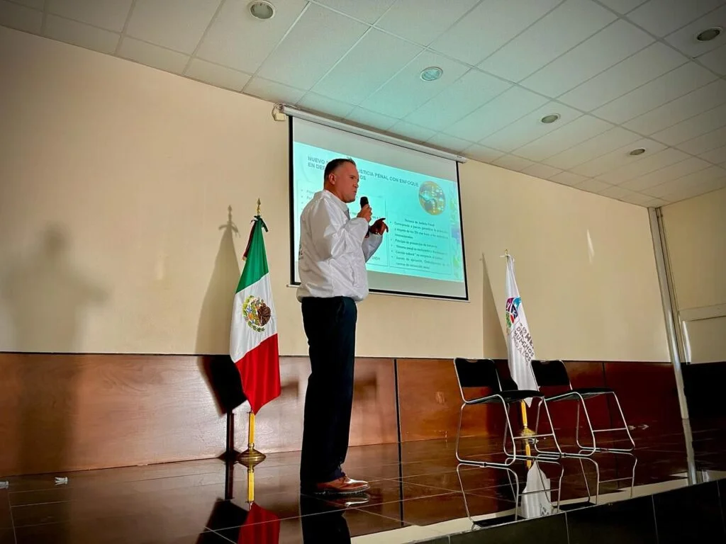 Escenario de un auditorio donde una persona de pie realiza una presentación utilizando un micrófono, frente a una pantalla con una diapositiva proyectada que incluye texto y gráficos. A un lado del escenario se encuentra la bandera de México y un estandarte institucional, mientras dos sillas vacías están colocadas detrás. El recinto cuenta con iluminación uniforme, paredes claras y techo con paneles.