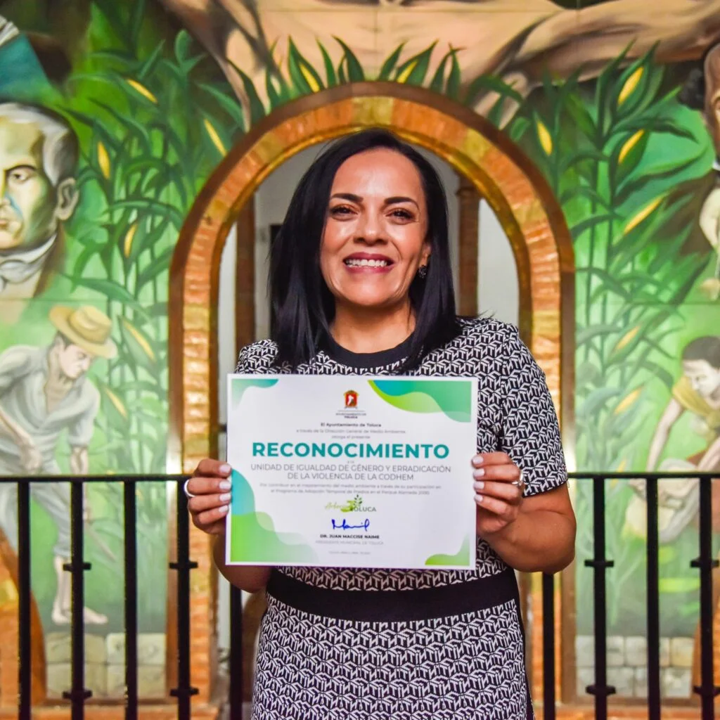 Persona de pie frente a un arco interior decorado con un mural colorido que representa figuras humanas y vegetación. La persona sostiene un certificado de “Reconocimiento” del Ayuntamiento de Toluca relacionado con una unidad de igualdad de género y erradicación de la violencia. El espacio cuenta con barandal metálico, iluminación uniforme y fondo artístico detallado.