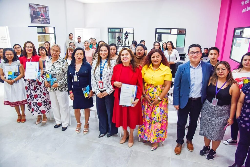 Grupo numeroso de personas posan de pie en una sala de capacitación con paredes blancas y una sección en color rosa. Varias personas sostienen revistas o documentos, mientras en el fondo se observan cámaras sobre tripiés y más asistentes. El espacio es amplio, con iluminación natural y mobiliario blanco.
