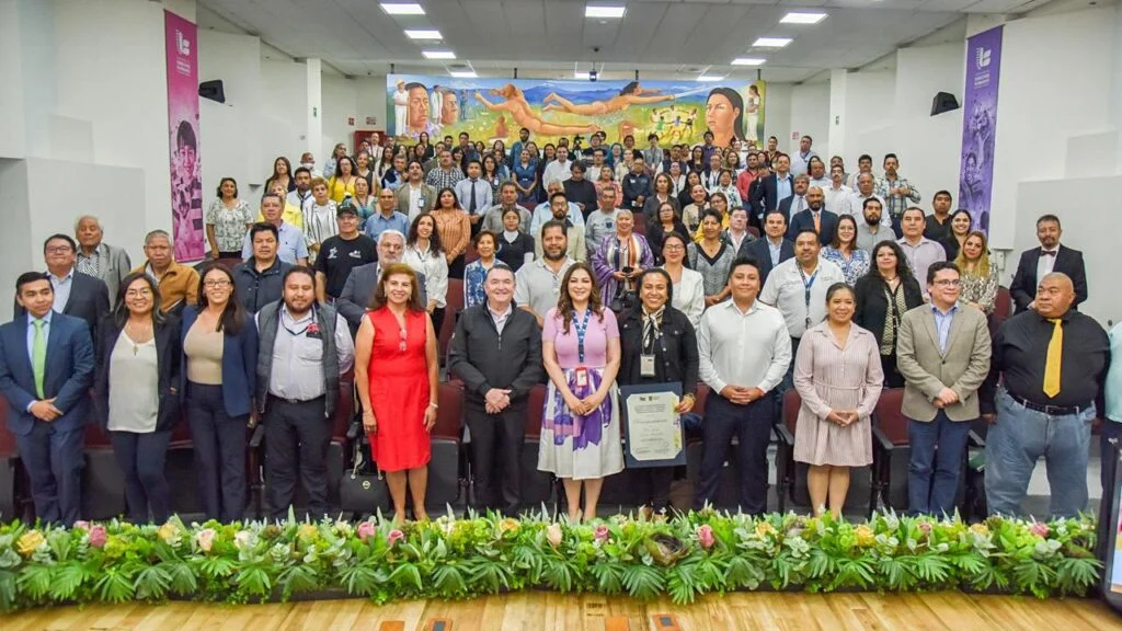 Fotografía grupal en un auditorio con decenas de asistentes organizados en filas, al frente de un escenario decorado con plantas; en la primera fila varias personas sostienen un documento y detrás se observa un mural colorido con figuras humanas en la pared.