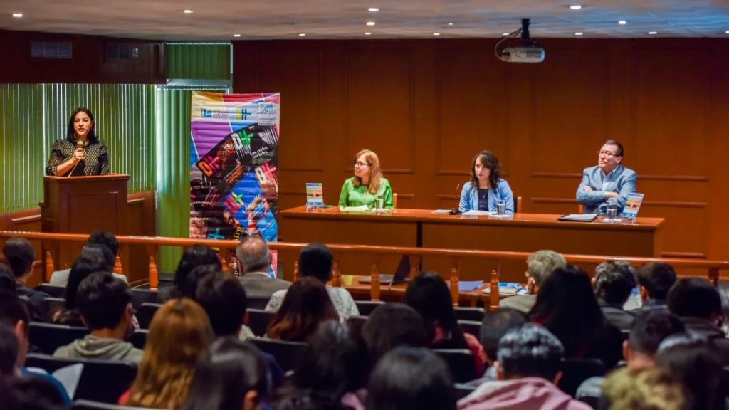 Vista general de una conferencia en auditorio, con una persona de pie hablando en un podio frente al público y tres personas participando en una mesa de panel al fondo; se observan asistentes sentados y un cartel informativo junto al escenario.
