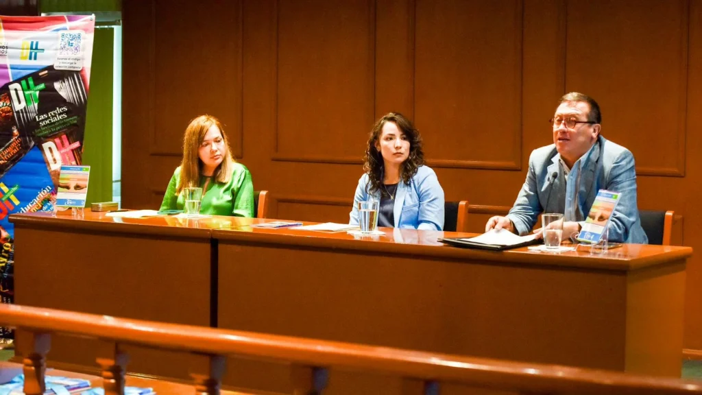 Tres personas sentadas detrás de una mesa en un panel dentro de un auditorio, con micrófonos, vasos de agua y material informativo sobre la mesa; al fondo se observa un panel de madera y un cartel vertical relacionado con derechos humanos.