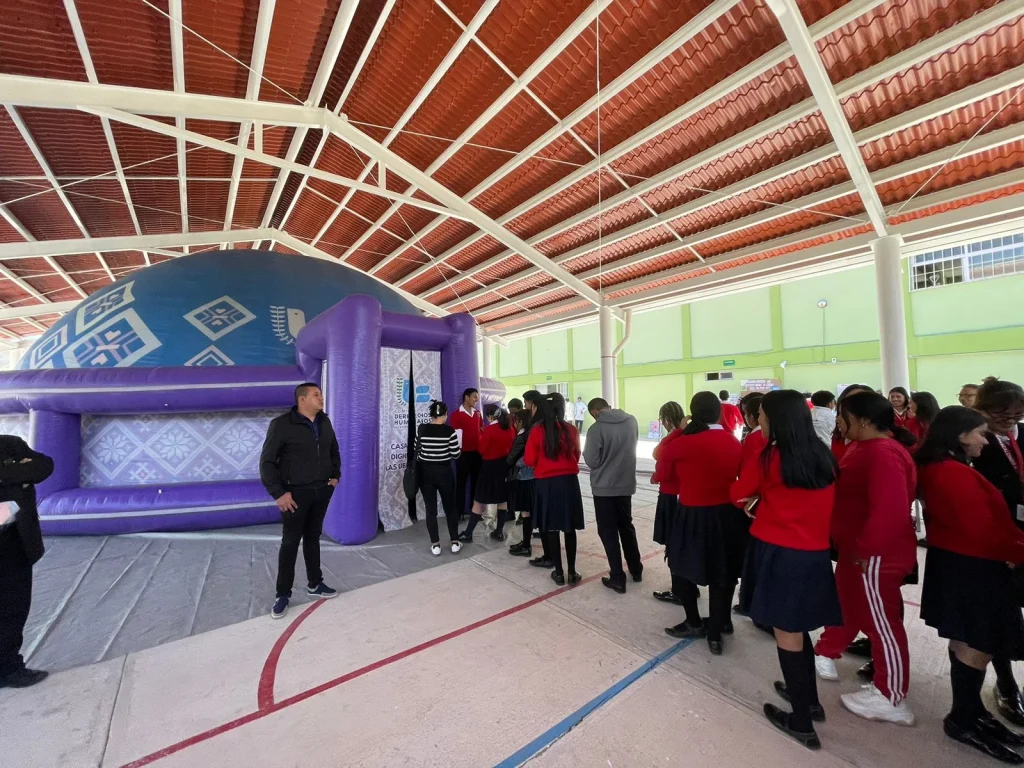 Grupo de estudiantes formados en fila dentro de un espacio techado con estructura metálica y techo rojo, esperando para ingresar a una estructura inflable morada con forma de domo; a un costado hay personal supervisando y paredes verdes con ventanas en el fondo.