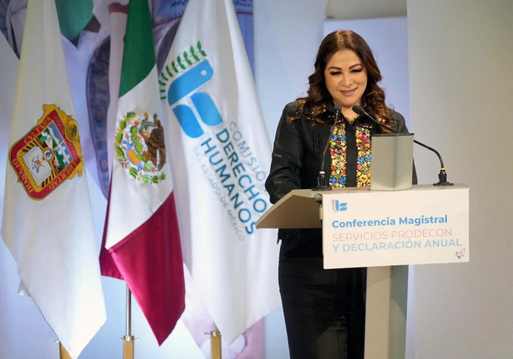 Persona exponiendo junto a un podio con micrófono durante una conferencia magistral, señalando hacia el público con banderas oficiales y logotipos de derechos humanos al fondo.