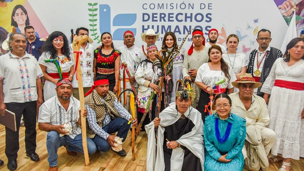 Fotografía grupal de varias personas vestidas con atuendos tradicionales y contemporáneos frente al logotipo de la Comisión de Derechos Humanos, posando en un auditorio interior.