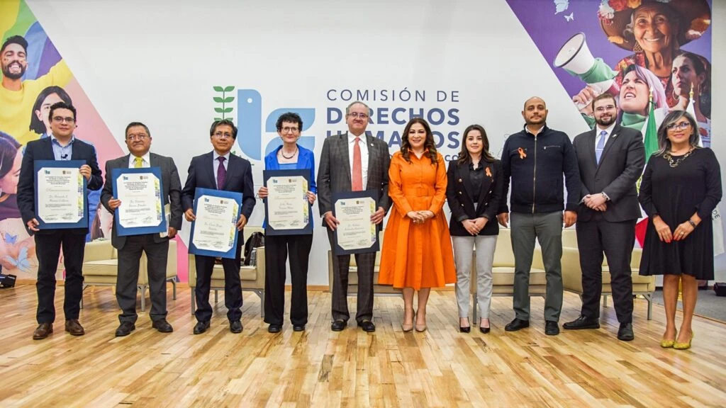 Grupo de personas posando en el escenario de un auditorio institucional, varias sostienen reconocimientos enmarcados, frente a un fondo con el logotipo de la Comisión de Derechos Humanos.