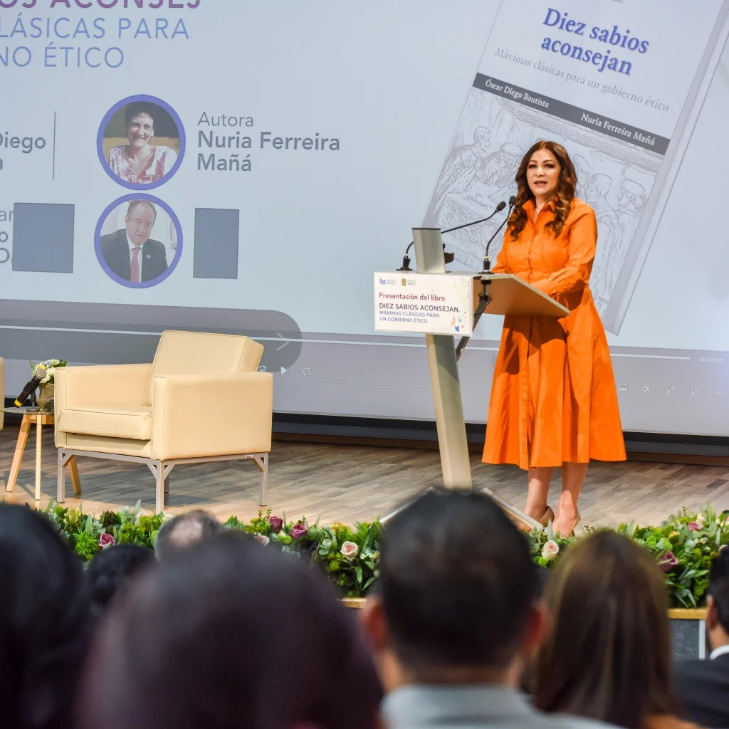 Persona de pie en un podio durante la presentación del libro “Diez sabios aconsejan”, en un auditorio, con una pantalla que muestra el nombre de la obra y la autora Nuria Ferreira Mañá.
