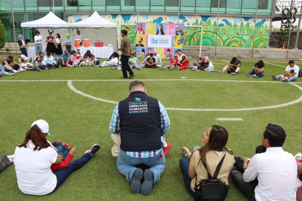 Personas sentadas formando un círculo en una cancha deportiva durante una actividad comunitaria organizada por la Comisión de Derechos Humanos, con carpas y materiales al fondo.