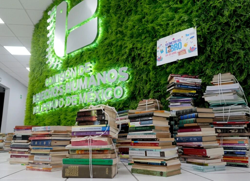 Pilas de libros apilados sobre el suelo frente a un muro verde decorativo con el logotipo iluminado de la Comisión de Derechos Humanos del Estado de México y un cartel del “Kilómetro del libro”.