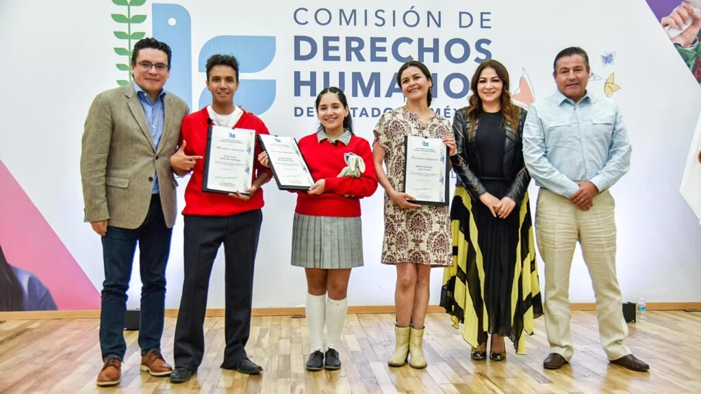 Grupo de personas en escenario institucional posando con reconocimientos enmarcados frente al logotipo de la Comisión de Derechos Humanos del Estado de México, durante una ceremonia de entrega.