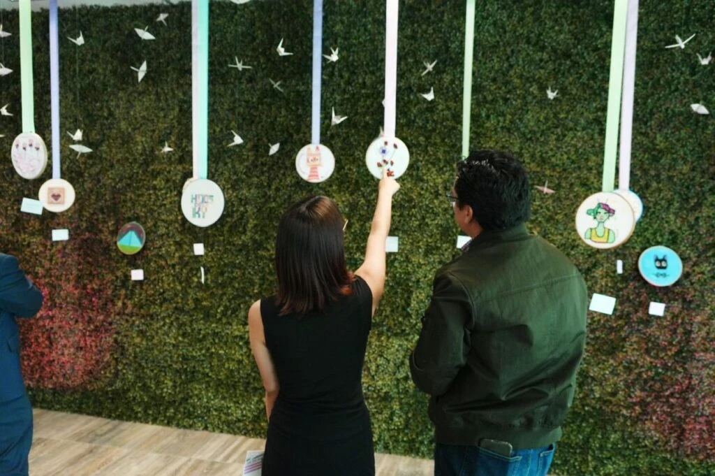 Dos personas observan una exhibición artística montada en un muro verde decorativo, donde diversas piezas circulares cuelgan de listones junto a figuras de papel en forma de aves.