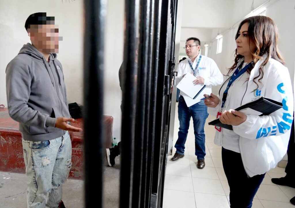 “Persona se encuentra detrás de una reja metálica mientras dos personas con portapapeles y documentos conversan con dicha persona en un pasillo interior con paredes claras.”
