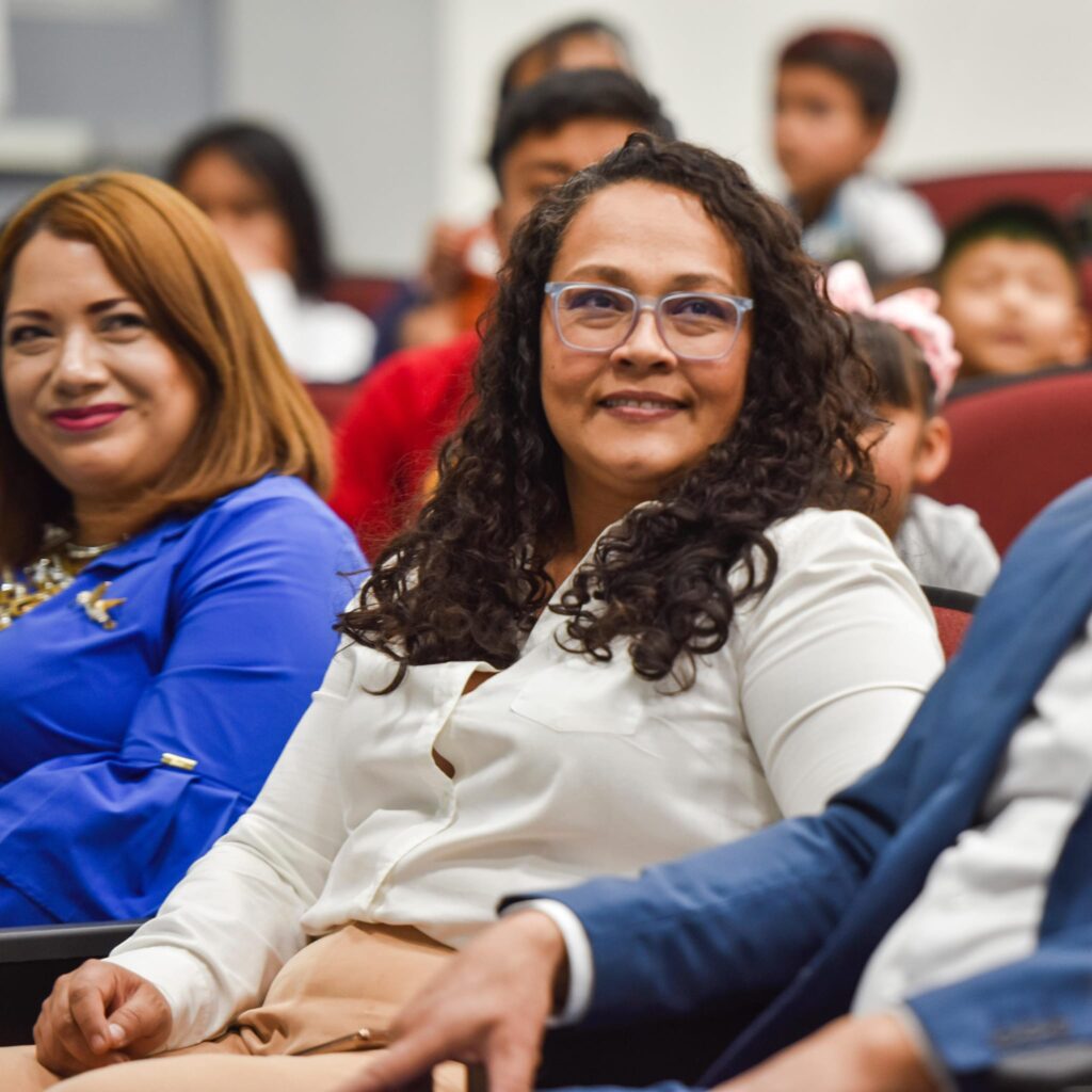 “Varias personas sentadas en un auditorio interior con butacas acolchonadas; en primer plano, una persona con gafas y blusa clara observa hacia un costado, mientras al fondo hay más asistentes, incluyendo menores, en un entorno de evento o reunión.”
