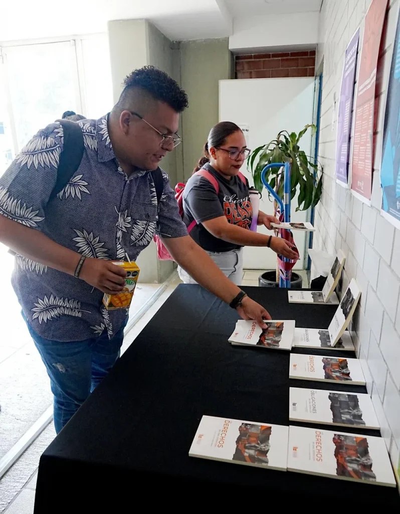 Dos personas observan y acomodan publicaciones sobre una mesa en un espacio interior, junto a un muro con carteles informativos.