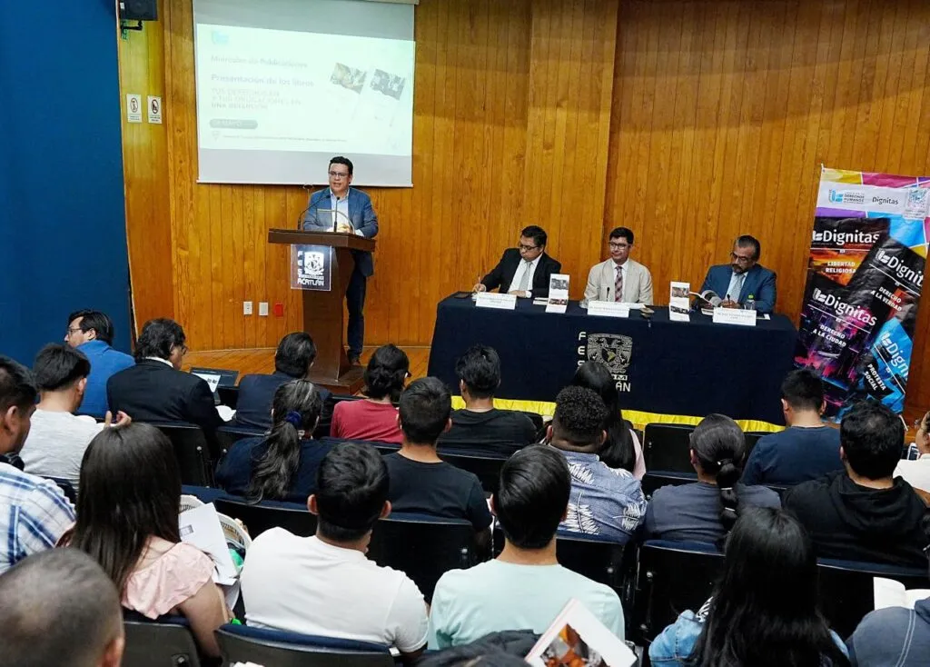 Auditorio con paredes de madera donde una persona expone desde un atril frente a una pantalla de proyección, mientras varias personas están sentadas como audiencia; a un lado, un panel de participantes se encuentra detrás de una mesa con libros y un cartel promocional visible.