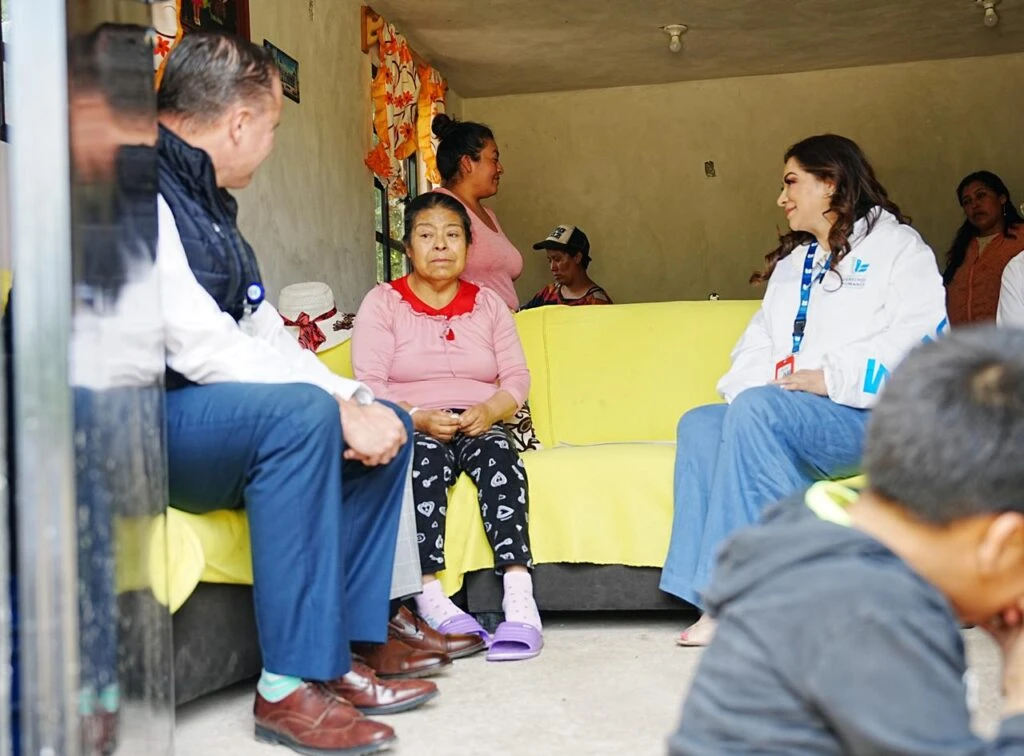 Varias personas reunidas en el interior de una vivienda, sentadas y de pie alrededor de un sofá amarillo mientras mantienen una conversación en grupo.