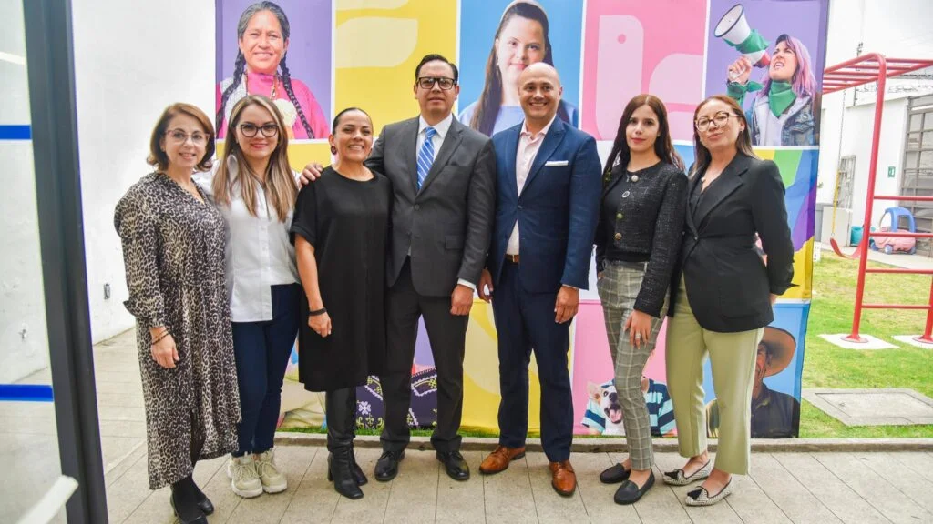 Grupo de personas posando de pie frente a un mural colorido con retratos, en un entorno exterior o semiabierto institucional.