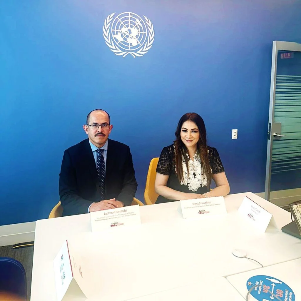 Dos personas sentadas en una mesa de trabajo con identificadores frente a una pared azul con el logotipo de las Naciones Unidas, en un entorno institucional.