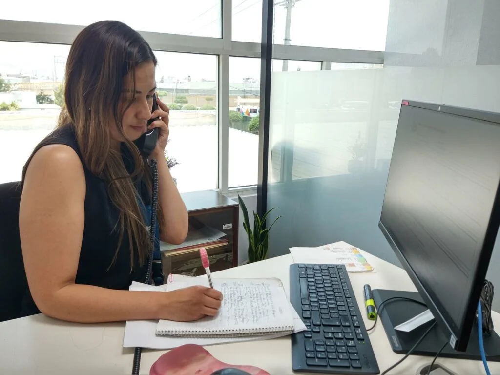 Persona trabajando en un escritorio, tomando notas en una libreta mientras habla por teléfono frente a una computadora en una oficina con vista exterior.