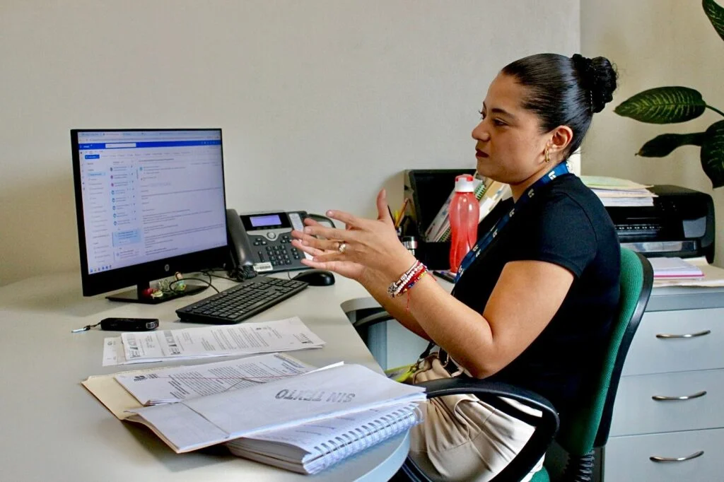 Persona sentada en una oficina frente a una computadora revisando información en pantalla y gesticulando mientras interactúa con documentos y un teléfono de escritorio.