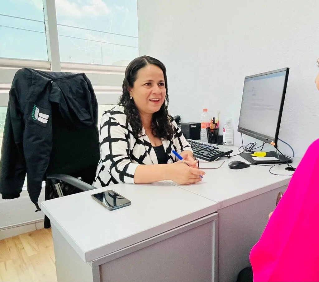 Persona sentada detrás de un escritorio en una oficina, interactuando con otra persona fuera de cuadro mientras sostiene un bolígrafo; sobre el escritorio hay una computadora, teléfono móvil y material de trabajo.