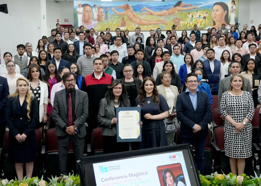 Grupo numeroso de personas posando en un auditorio tras un evento, con una persona al frente sosteniendo un reconocimiento y un mural artístico visible en la pared posterior.