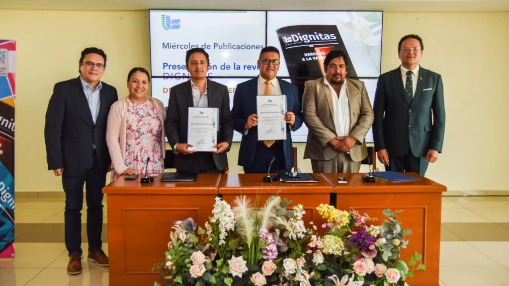 Grupo de personas en una mesa principal durante la presentación de una publicación titulada “Dignitas”, sosteniendo ejemplares y documentos frente a una pantalla institucional.