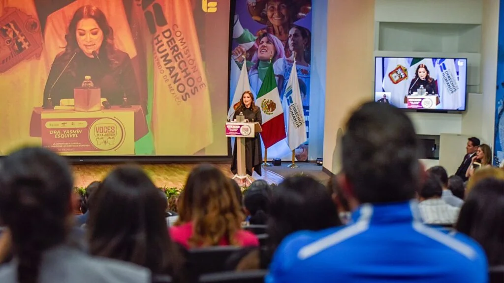 Vista desde el público de un auditorio donde una persona ofrece un discurso en el escenario; una pantalla grande proyecta la misma imagen del podio y se observan banderas institucionales junto al escenario.
