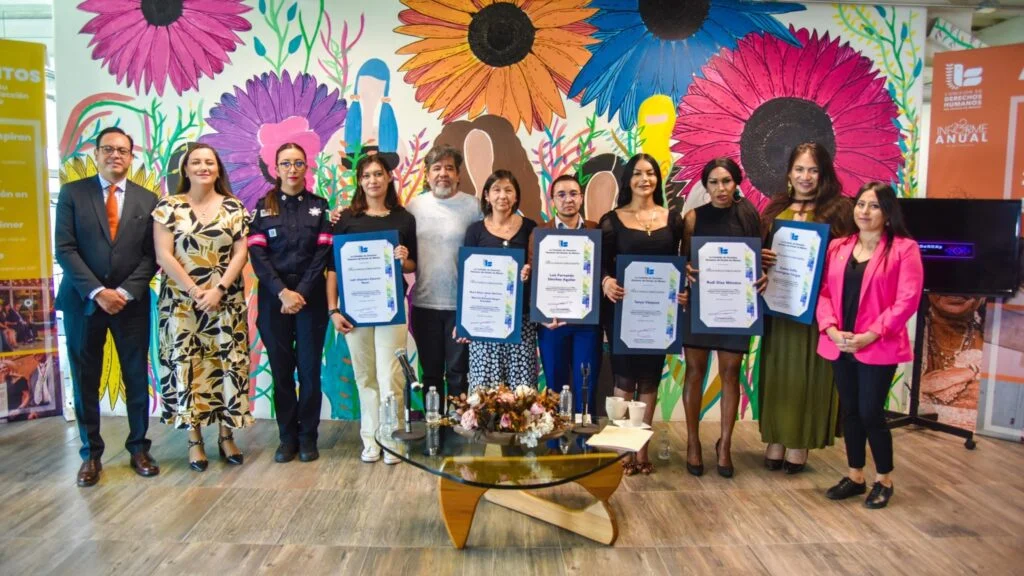 Grupo de personas posando en fila dentro de un espacio interior, varias sostienen reconocimientos impresos; al fondo hay un mural colorido con flores grandes y una mesa de centro con arreglo floral en primer plano.