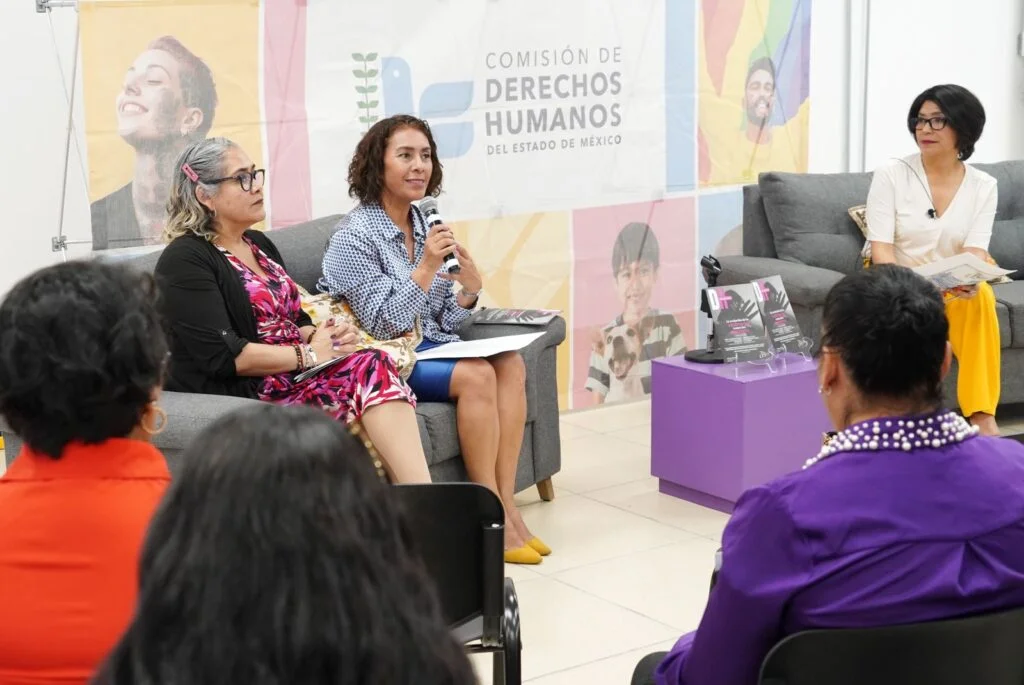 Tres personas participan en un panel de diálogo; una de ellas sostiene un micrófono mientras habla, con público sentado al frente y material informativo sobre una mesa lateral.