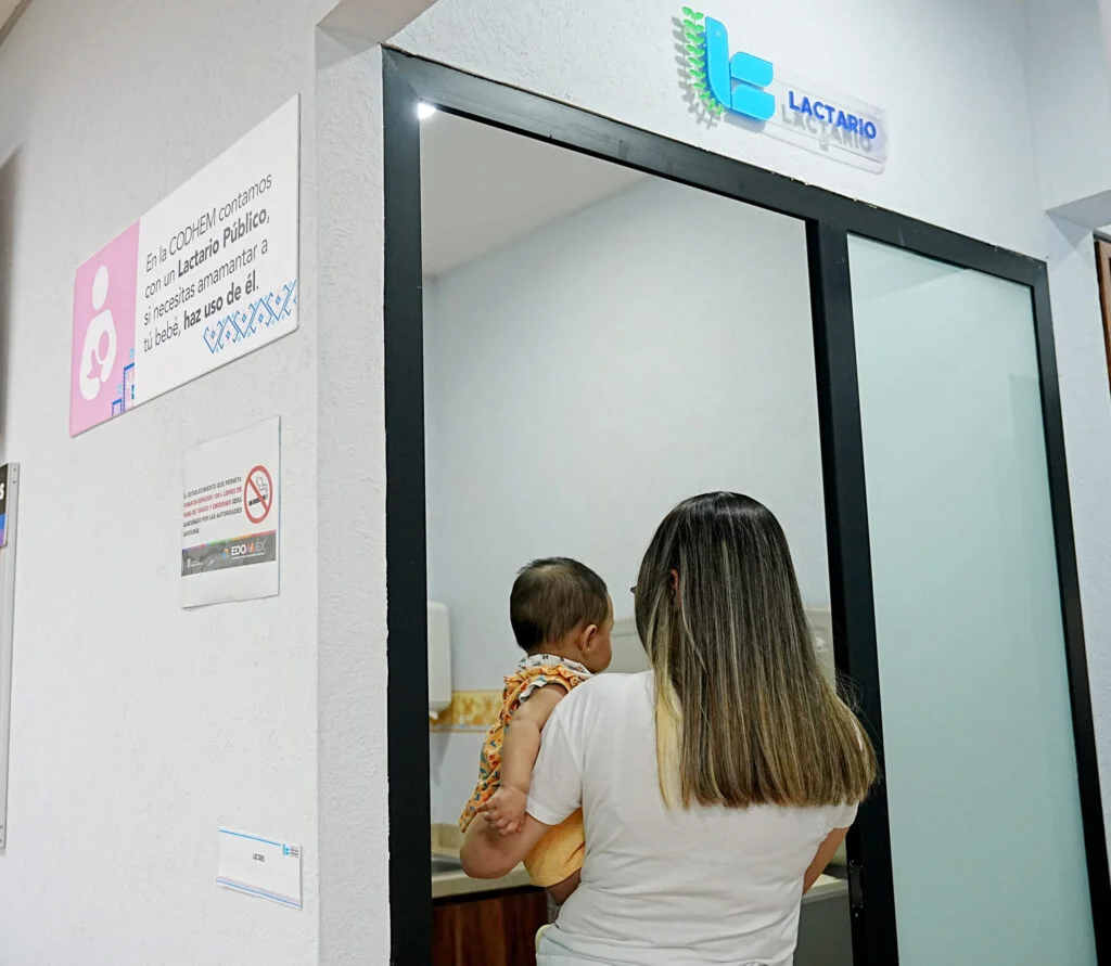 Acceso a un lactario en un edificio institucional; una persona ingresa cargando a un bebé, junto a un letrero que indica el uso del espacio para lactancia y el logotipo institucional colocado sobre la puerta.