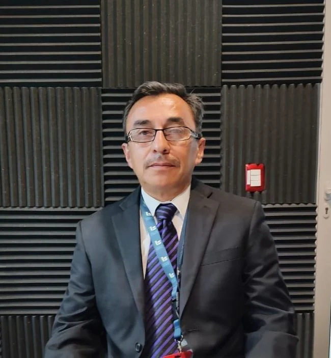 Persona con vestimenta formal, traje oscuro, camisa clara y corbata, sentada frente a un fondo con paneles acústicos; porta gafas y un gafete colgante, en un entorno que parece ser un estudio o sala de grabación.