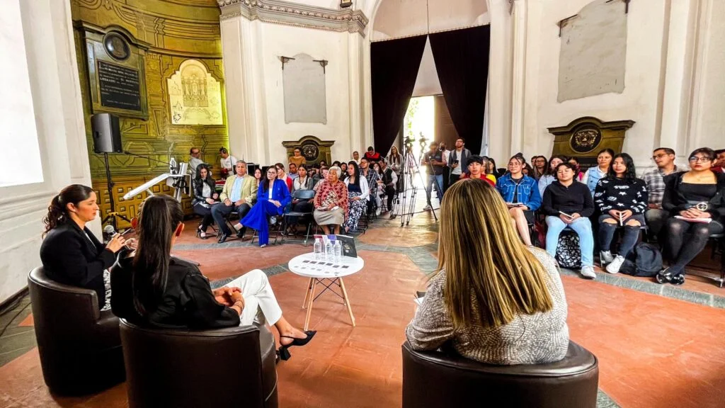 Vista frontal de una charla en un recinto histórico; varias personas están sentadas frente a tres participantes ubicados en sillones alrededor de una mesa, mientras una persona utiliza un micrófono para intervenir durante la conversación.