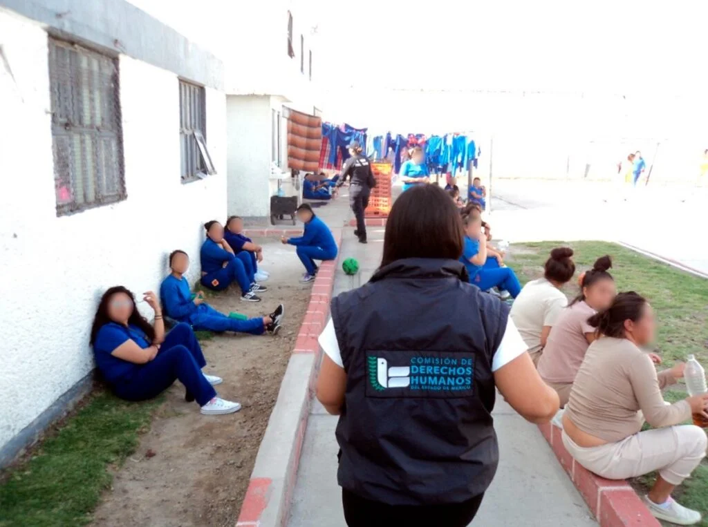 Espacio exterior dentro de una instalación donde varias personas se encuentran sentadas o caminando; en primer plano, una persona con chaleco de la Comisión de Derechos Humanos se dirige hacia un grupo reunido junto a un muro.